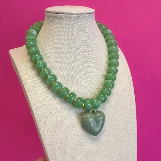 A green beaded necklace with a carved green heart pendant, displayed on a beige jewelry bust against a bright pink background. A hand is visible holding the side of the bust.