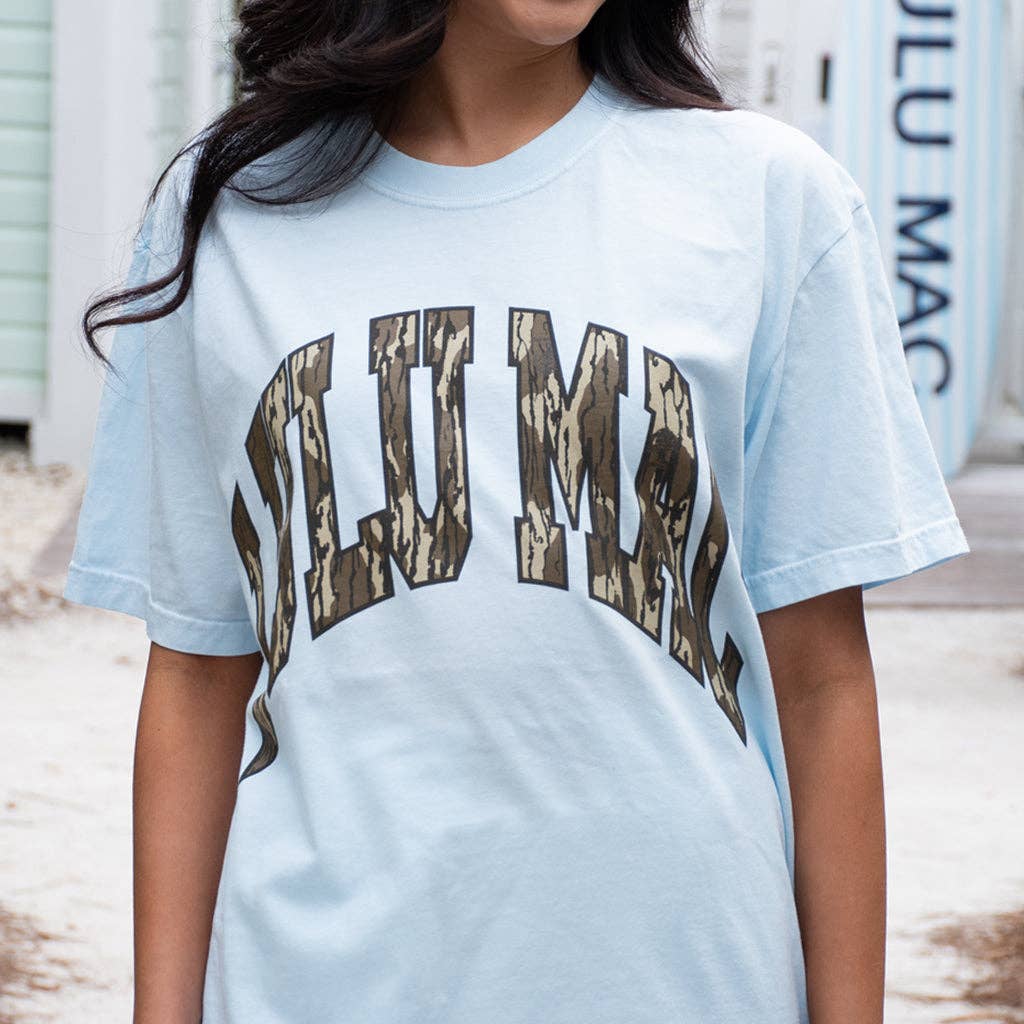 Woman wearing Lulu Mac Mossy Oak Bottomland® Comfort Colors® Graphic Tee featuring Mossy Oak camouflage lettering on a chambray blue shirt.