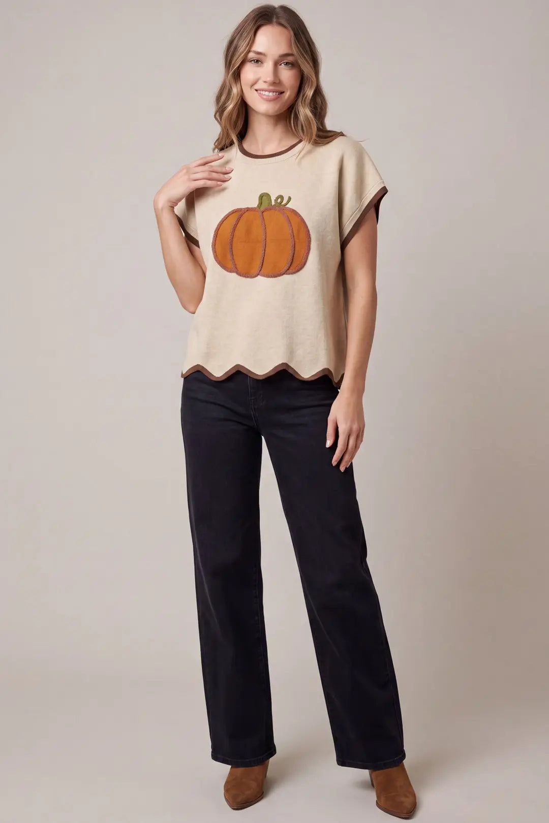 A woman modeling a beige knit top with a large orange pumpkin graphic on the front, paired with dark wide-leg pants and brown ankle boots, against a solid beige studio background.