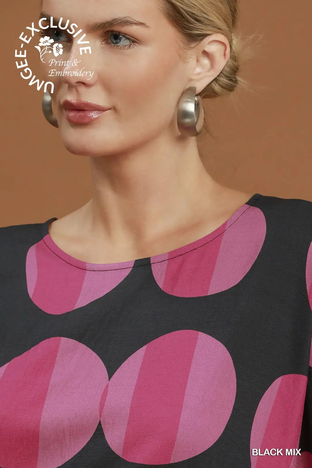 Close-up of a woman wearing a black top with large pink and magenta wavy circular patterns, paired with chunky silver hoop earrings, against a warm brown background. A watermark for 'Unbe-F Exclusive Print & Embroidery' is visible in the top left.