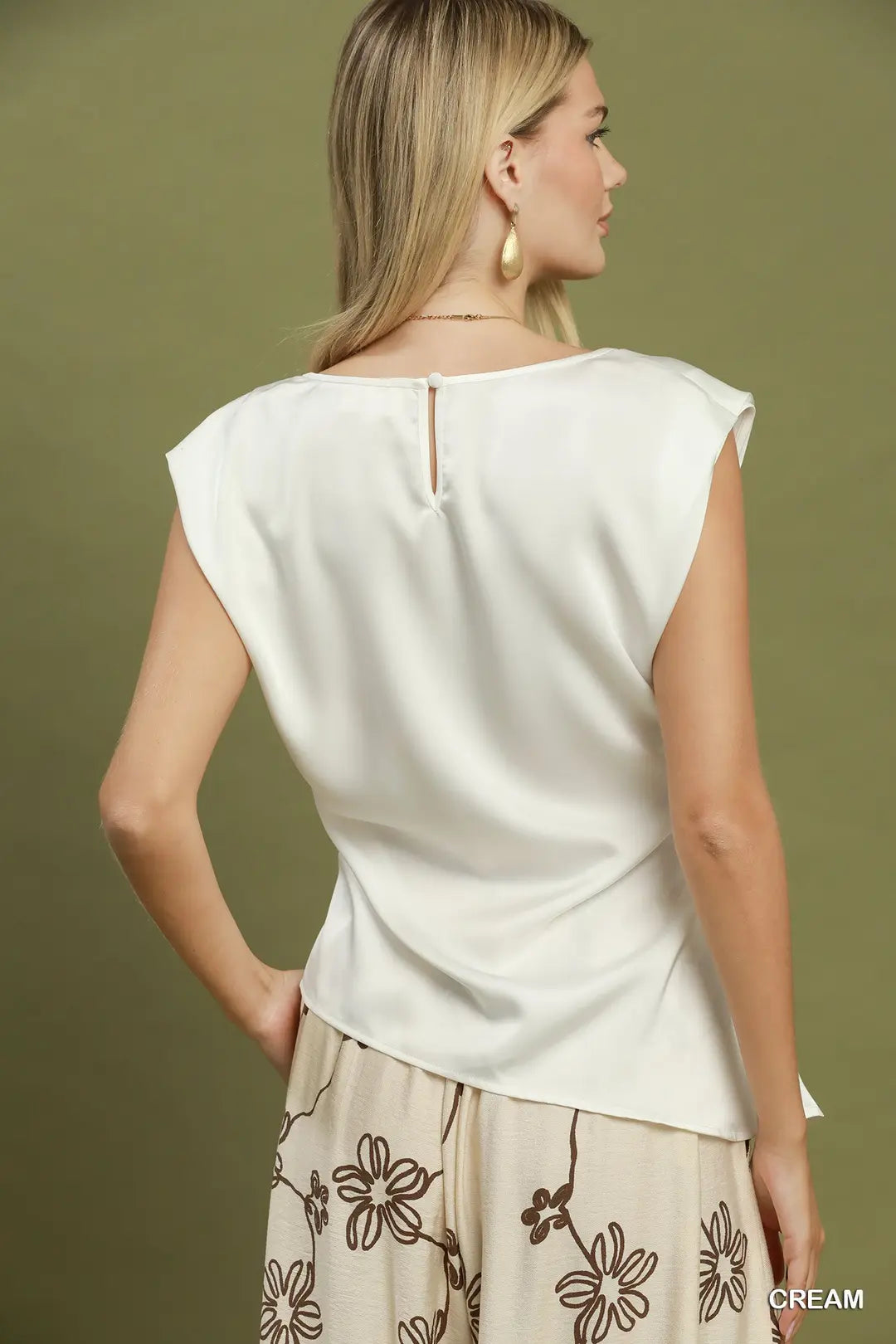 A woman shown from the back wearing a cream satin-like sleeveless top with a small keyhole cutout at the neckline, paired with beige floral pants against an olive green background.