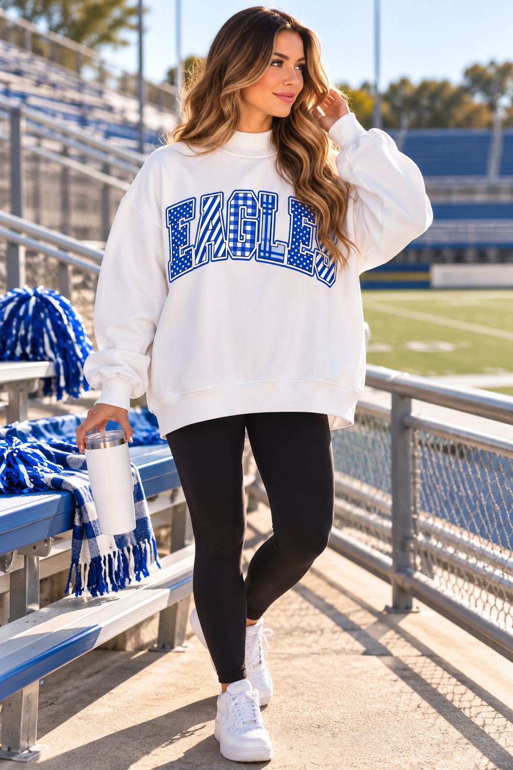 A woman stands on a stadium walkway wearing a white sweatshirt with a blue patterned 'EAGLE' graphic across the chest, black leggings, and white sneakers. Blue and white pom-poms and blankets are visible on the bleachers behind her.