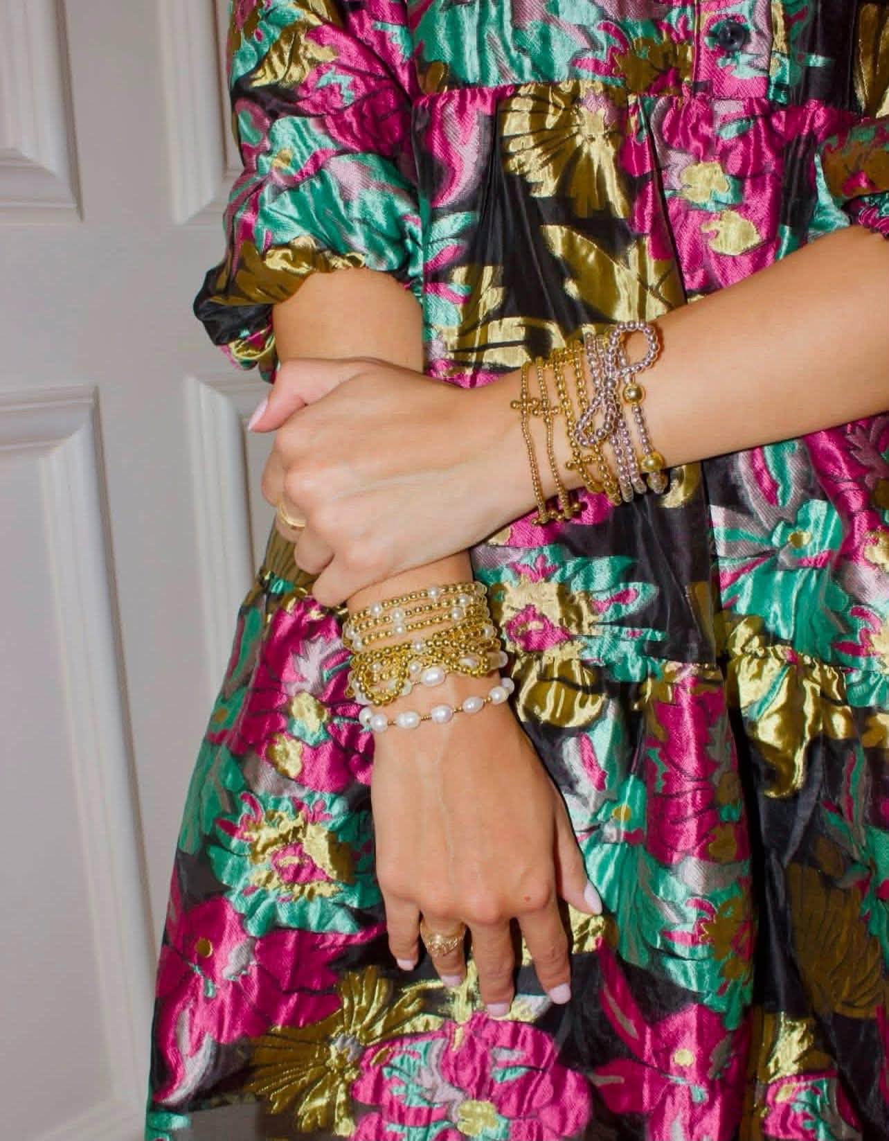 A close-up of a person wearing a vibrant floral dress with pink, green, and gold patterns, accessorized with multiple gold and pearl bracelets on both wrists.
