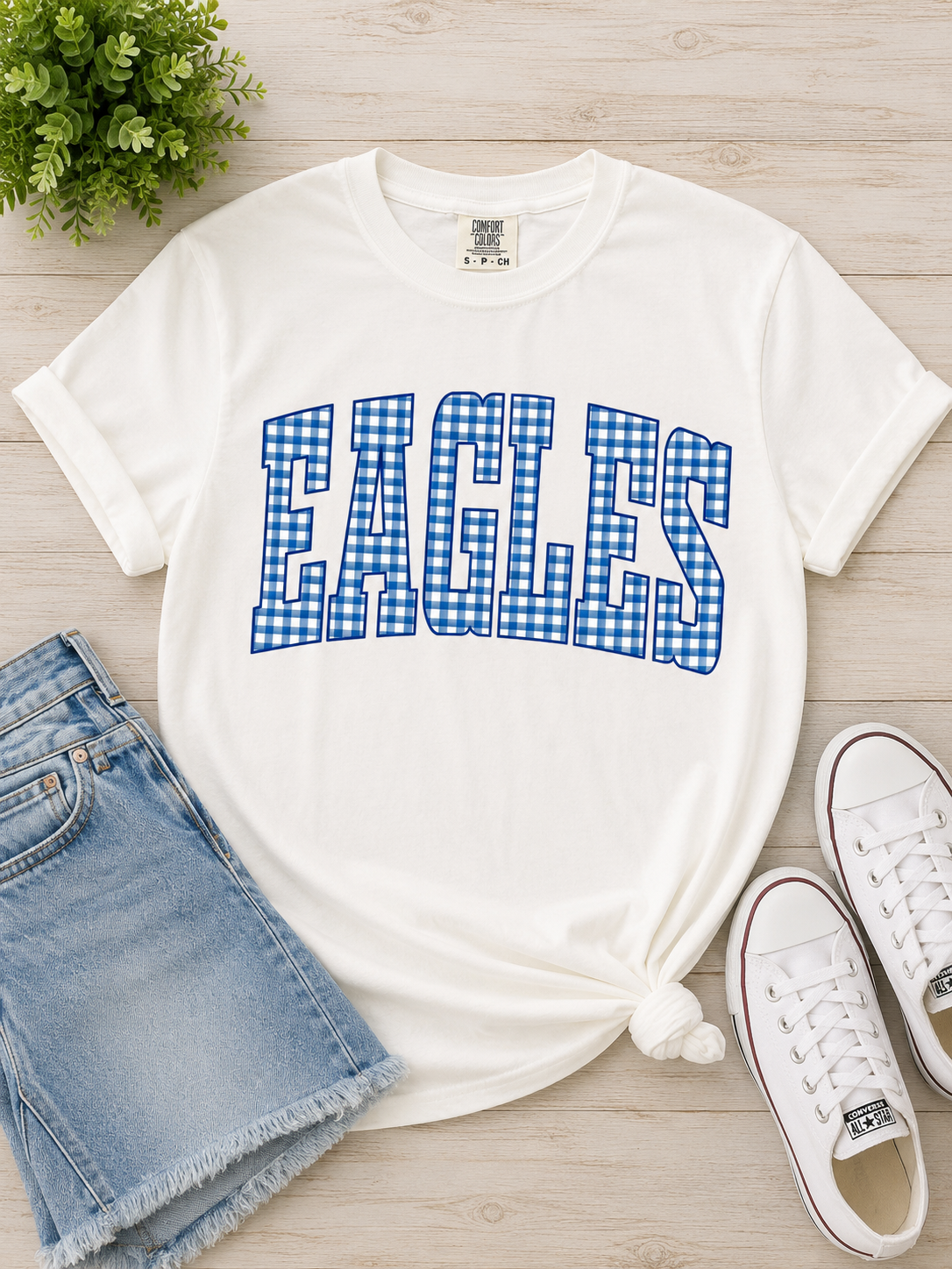 A white short-sleeve graphic T-shirt laid flat on a light wood surface, styled with denim shorts, white sneakers, and a small green plant. The shirt has large 'EAGLES' lettering across the chest in a blue-and-white gingham pattern.