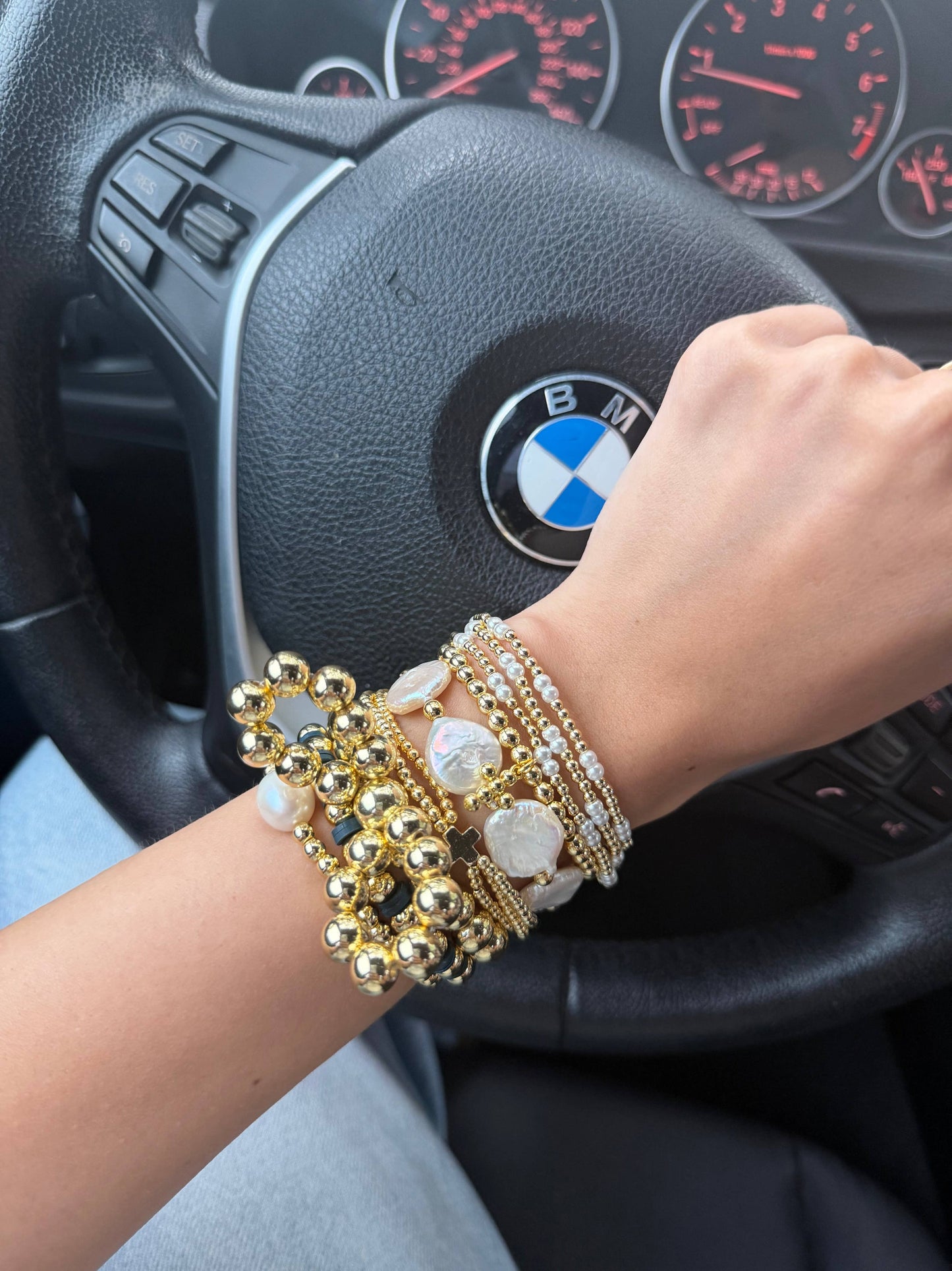 A close-up of a person's wrist wearing a stack of gold and pearl bracelets, resting on a car steering wheel with a visible BMW logo.