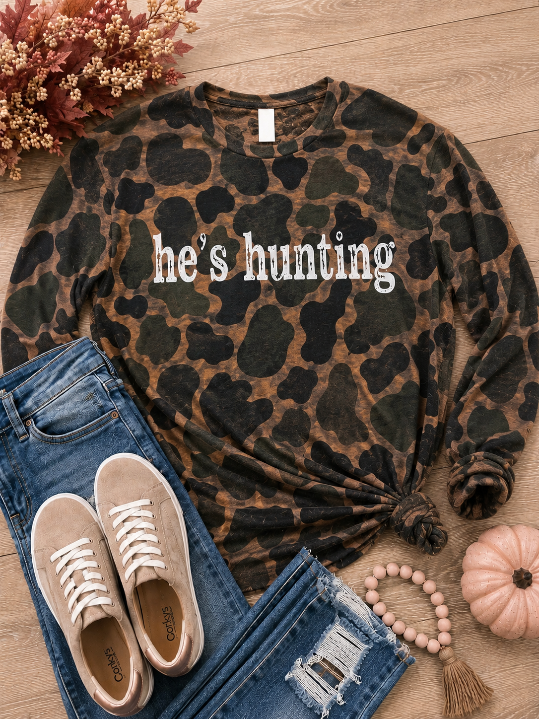 A long-sleeve crewneck sweatshirt with a dark green and brown animal print pattern and the text 'he's hunting' printed across the chest. The sweatshirt is styled in a flat lay with blue jeans, beige sneakers, a pink beaded tassel necklace, a small pumpkin, and autumn foliage on a light wood background.