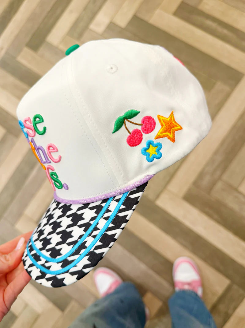 White cap with colorful designs and text held by a hand on a wooden floor.