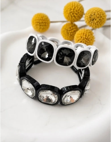 Two stretch bracelets displayed on a white marble surface with yellow decorative pom-pom flowers in the background. One bracelet is black with clear faceted rhinestones, and the other is white with black faceted rhinestones.