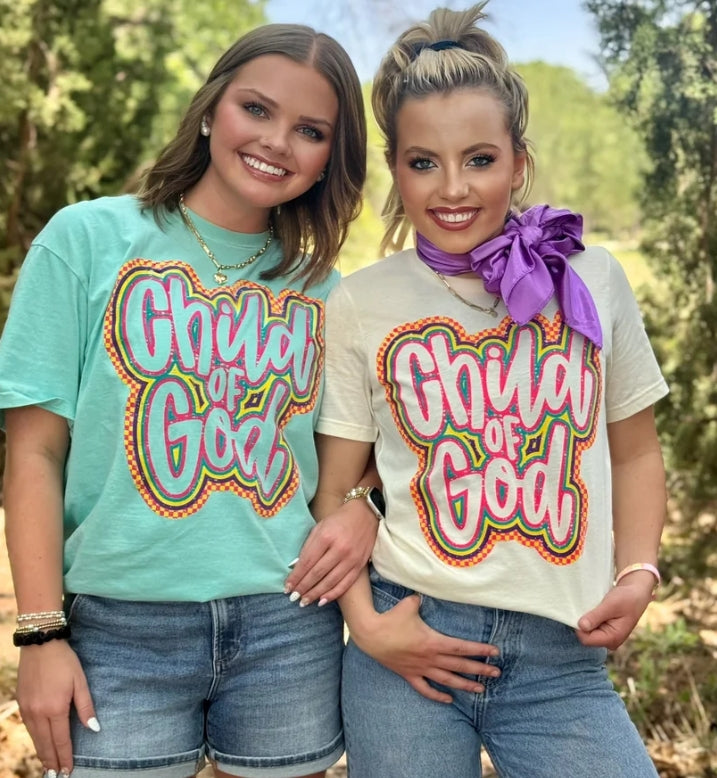Two women standing outdoors in front of trees, wearing matching graphic T-shirts with the phrase 'Child of God' in large pink and white lettering with orange, yellow, and green outline details. The woman on the left wears a mint green shirt, and the woman on the right wears an off-white shirt with a large purple bow around her neck.