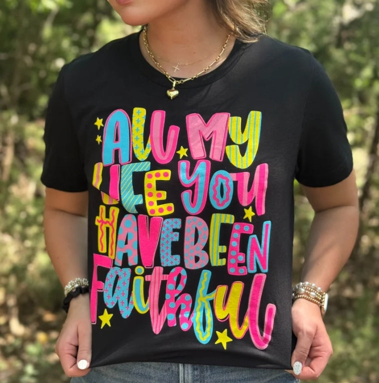 A woman wearing a black t-shirt with a colorful graphic that reads 'ALL MY LIFE YOU HAVE BEEN Faithful' in pink, blue, yellow, and green letters, set against a blurred outdoor background.