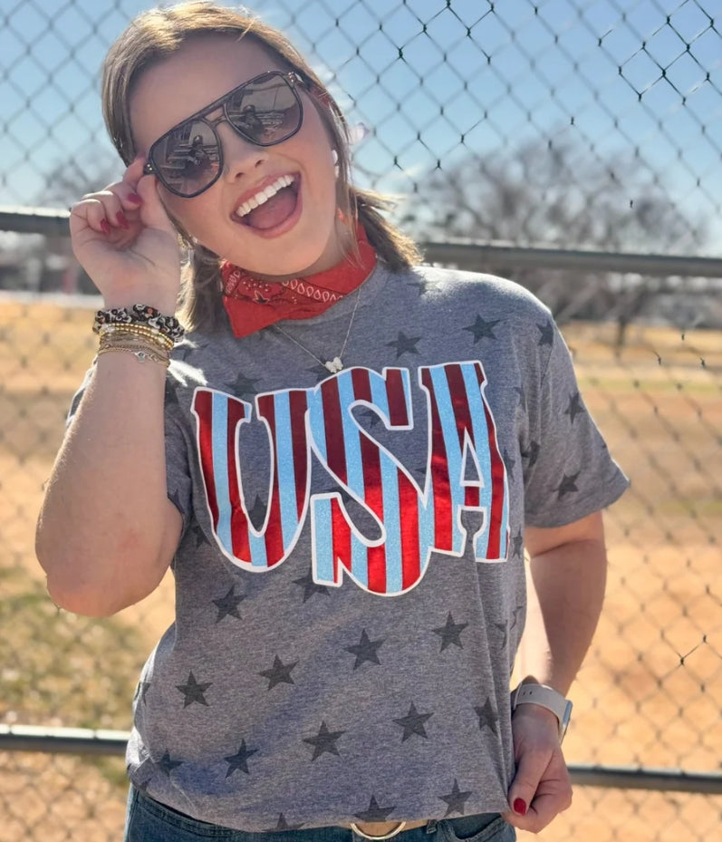 Woman wearing a gray 'USA' t-shirt with red, white, and blue colors, standing in front of a chain-link fence.