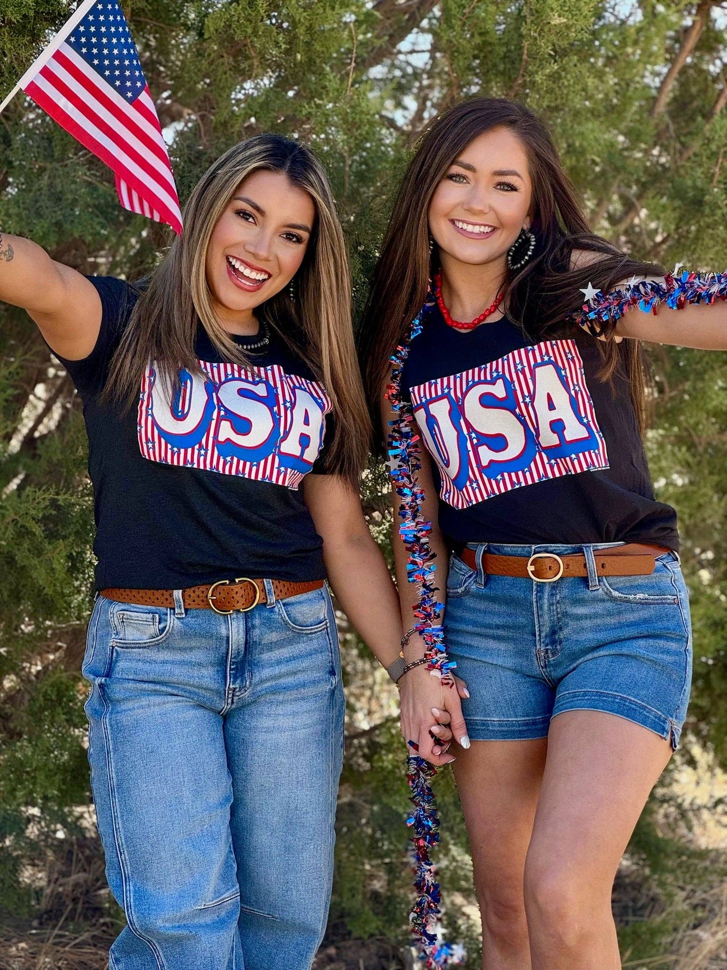 Two women wearing 'USA' shirts with an American flag, standing outdoors with trees in the background.