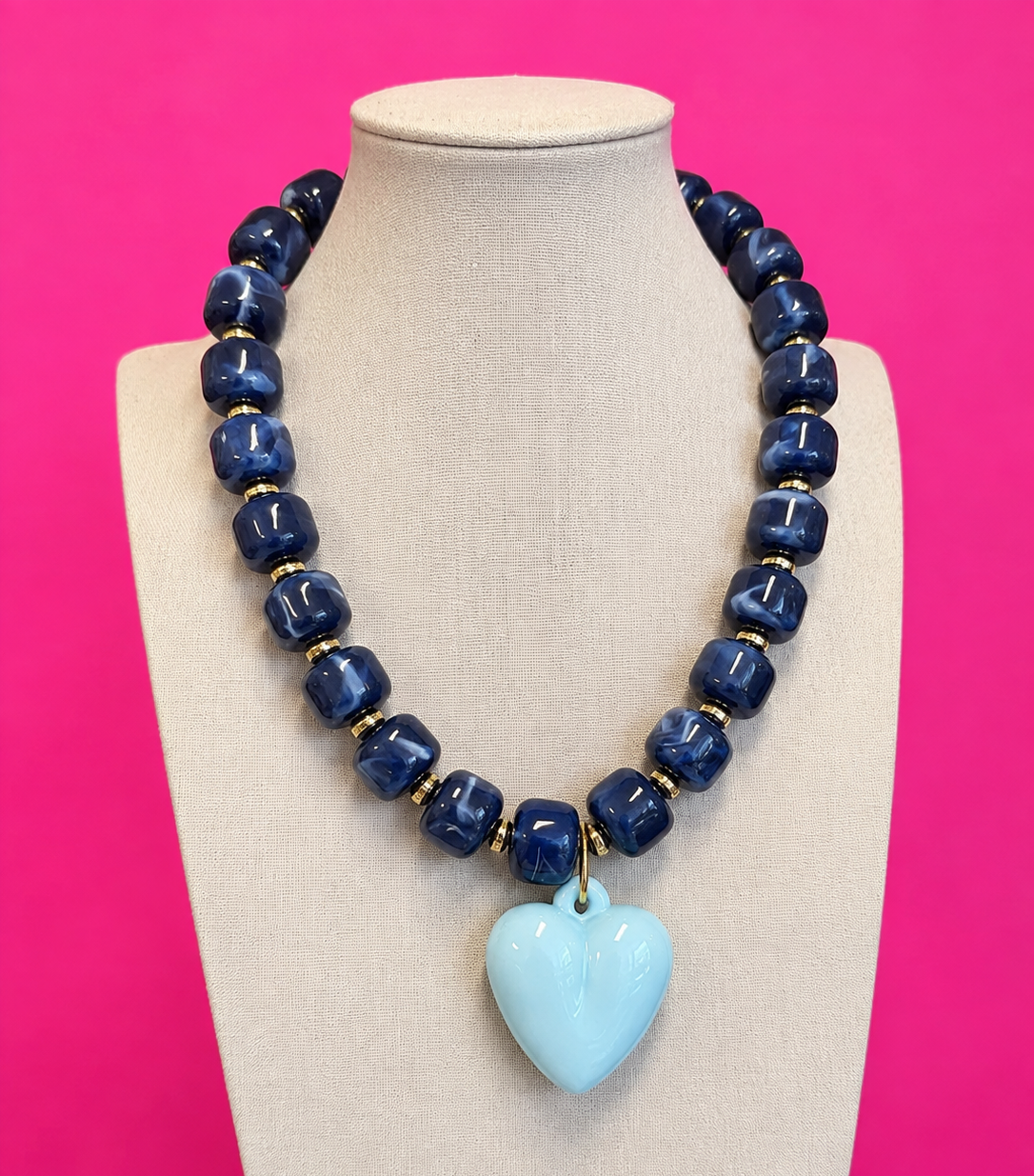 A beaded necklace displayed on a beige fabric mannequin bust against a bright pink background. The necklace features dark blue faceted beads separated by small gold spacer rings, with a light blue heart-shaped pendant at the center.