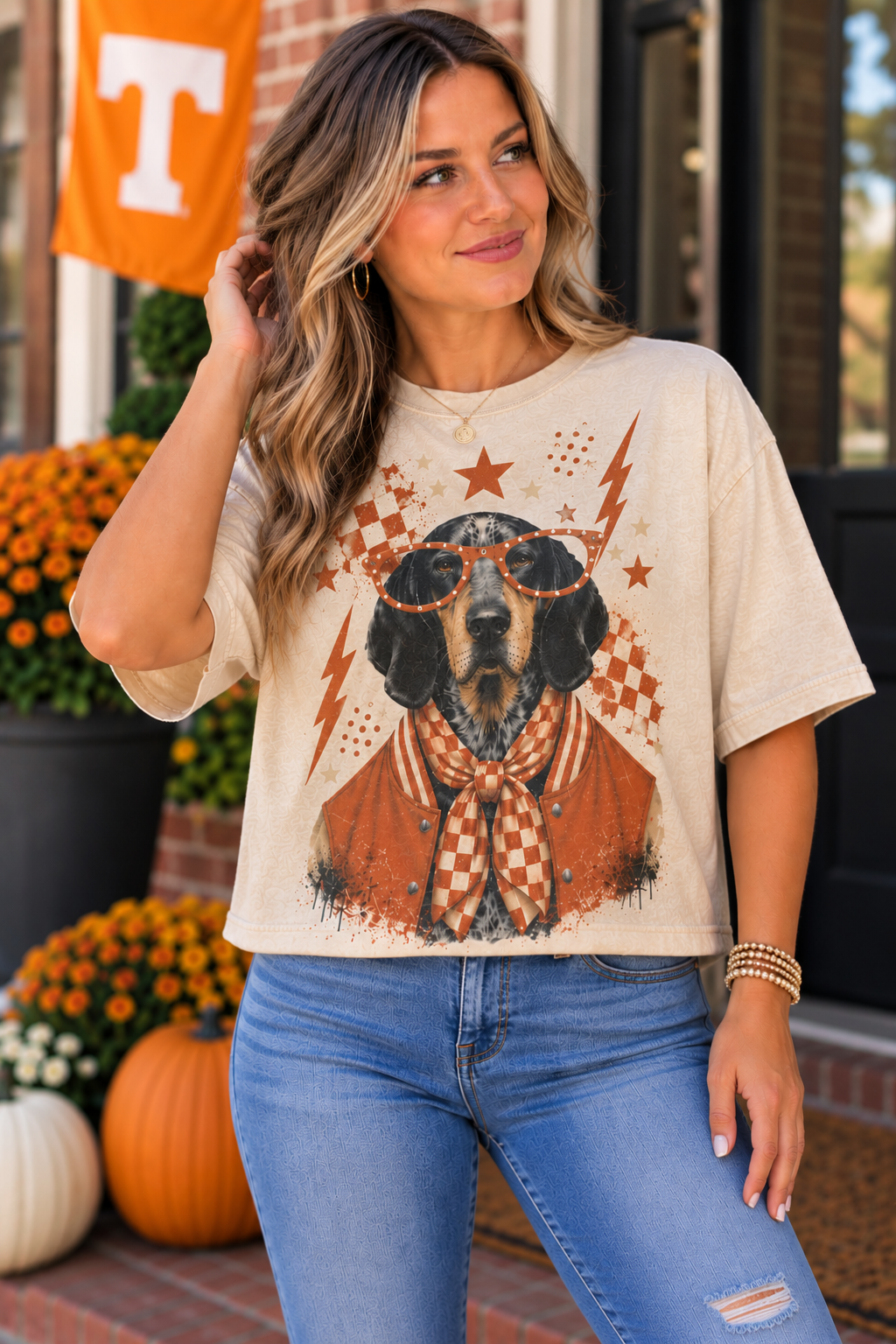 A woman wearing a beige graphic t-shirt with a dachshund wearing sunglasses and an orange-and-white checkered bow tie, paired with blue jeans in a fall-themed outdoor setting with pumpkins and an orange Tennessee flag.