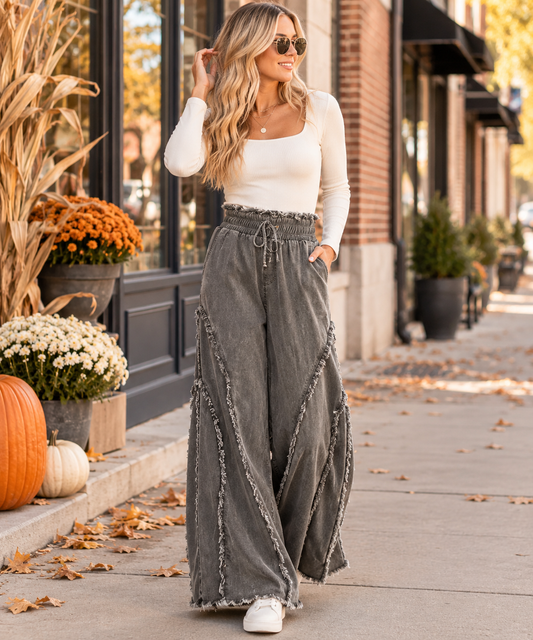 A woman stands on a city sidewalk wearing a white long-sleeve top and high-waisted, wide-leg gray denim pants with fringe trim. The background shows a storefront with autumn decorations including pumpkins, mums, and corn stalks.