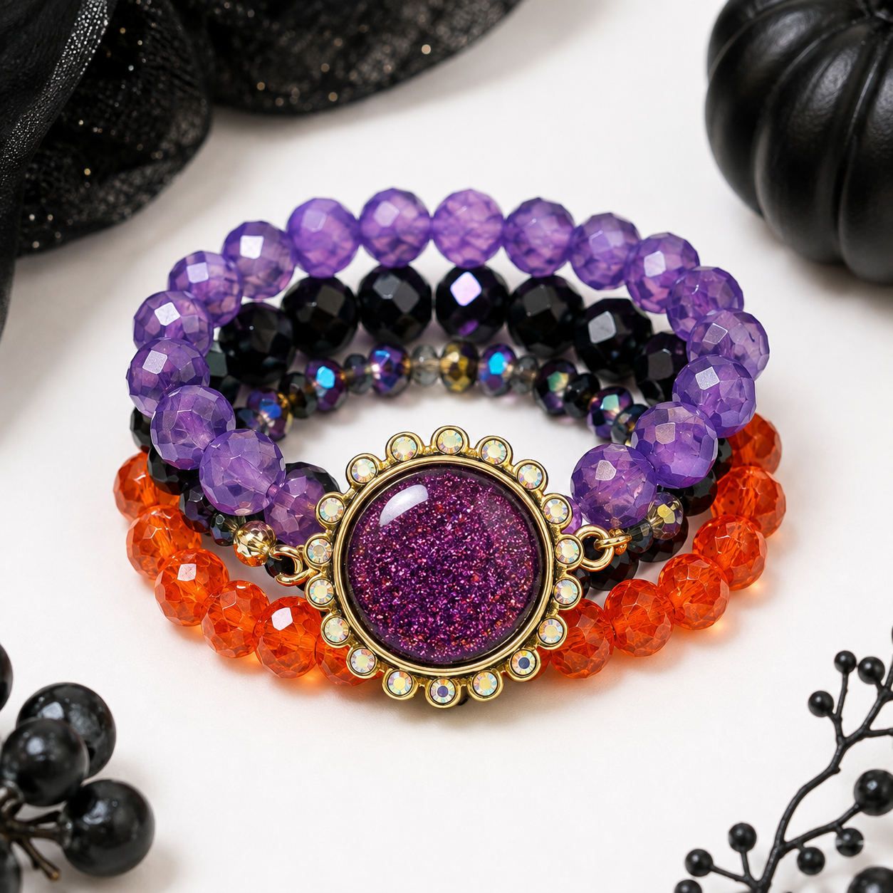 A set of three beaded stretch bracelets stacked together on a white surface. The top bracelet is made of faceted purple beads, the middle is made of faceted black beads, and the bottom is made of faceted orange beads.