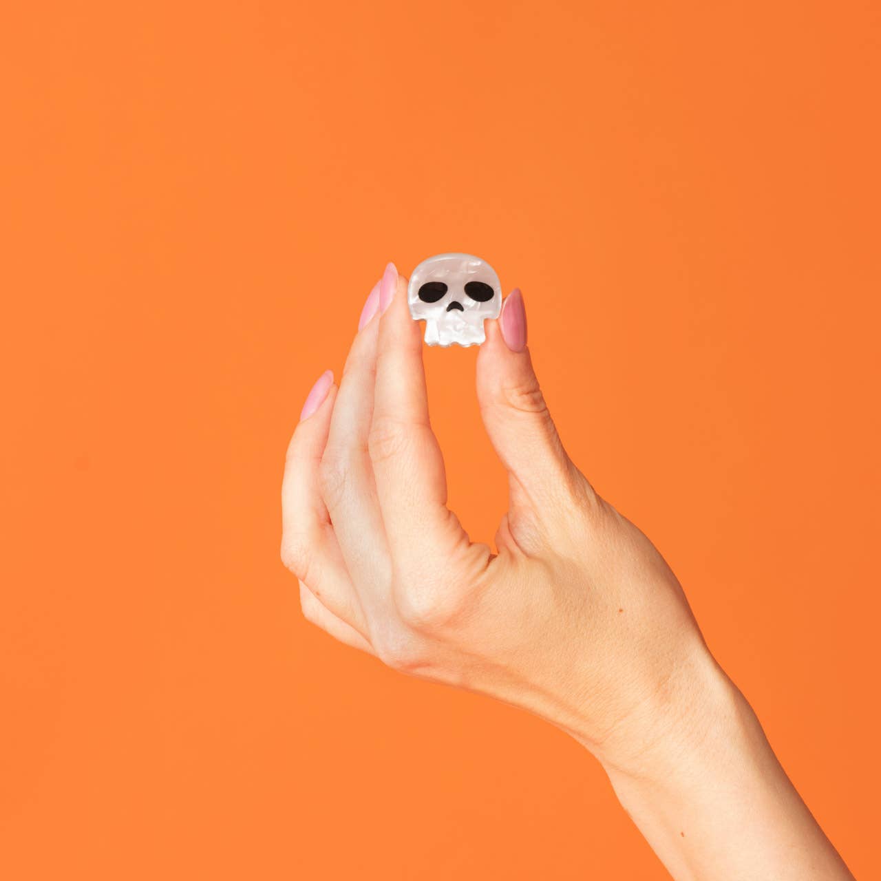 A hand with pink manicured nails holds a small white skull-shaped ring against a solid orange background.