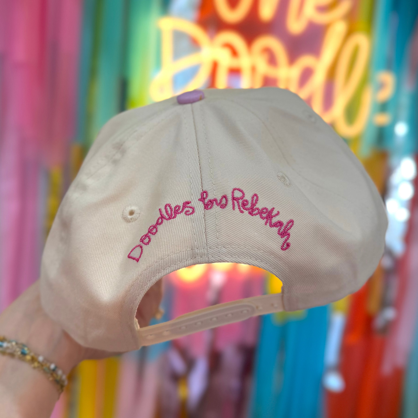 Beige cap with 'Doodles for Rebekah' text held in front of a colorful neon sign.