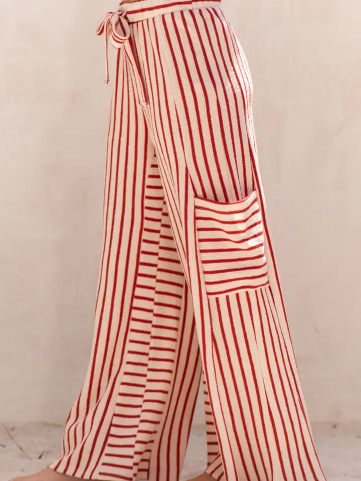Red and off-white vertically striped wide-leg pants with a tie waist and a large patch pocket on the left leg, shown on a model against a neutral beige background.