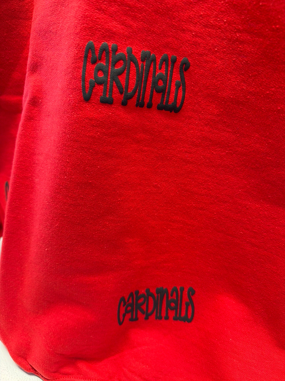 A close-up of a bright red fabric item, likely a sweatshirt or hoodie, featuring the word 'Cardinals' embroidered in black across the chest and again near the lower front.