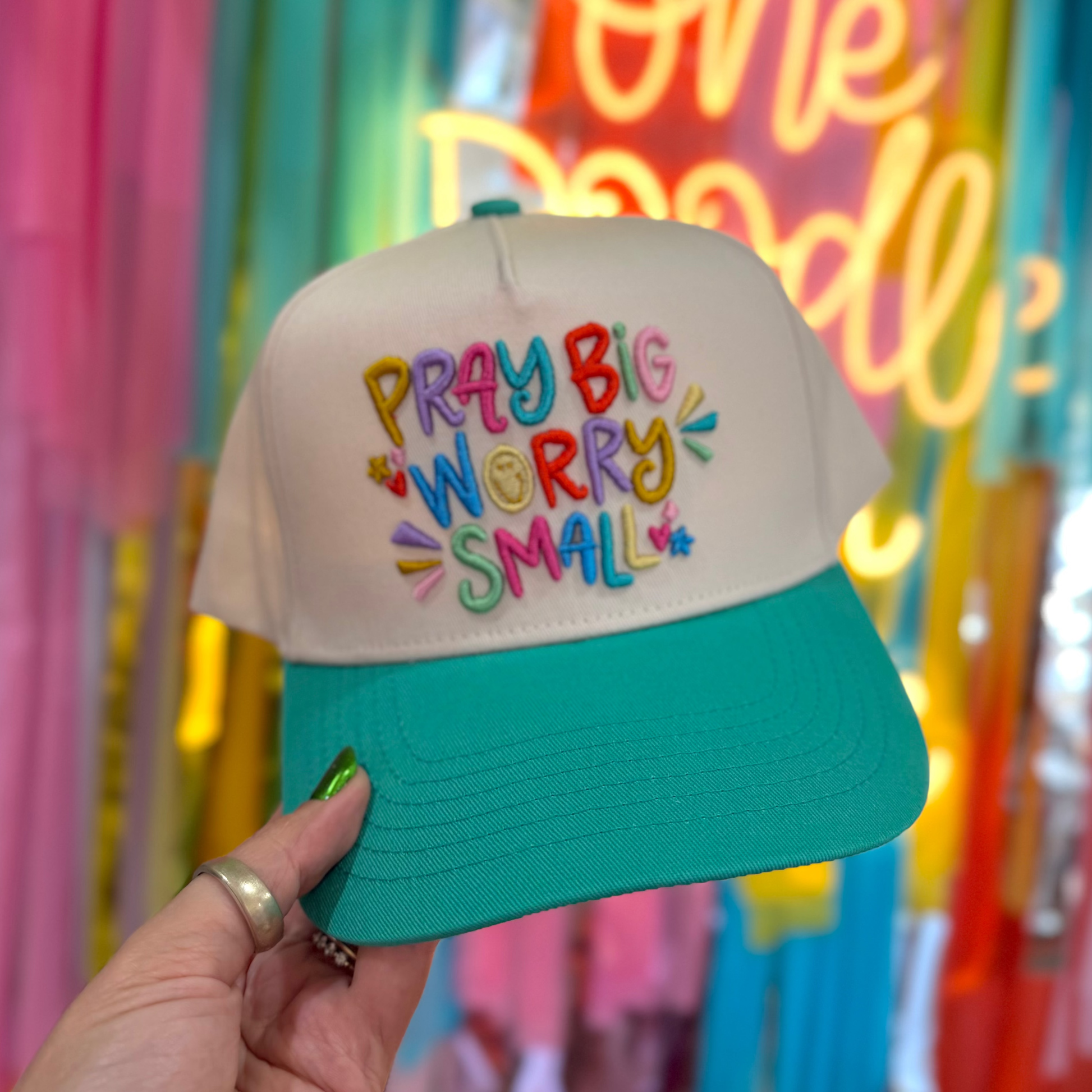 Cap with 'Pray Big Worry Small' text held in front of colorful neon lights