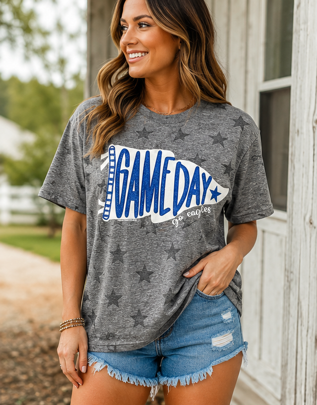 A woman wearing a heather gray graphic t-shirt with a football-shaped 'GAMEDAY' design in blue and white, paired with denim shorts, standing outdoors next to a white wooden door.