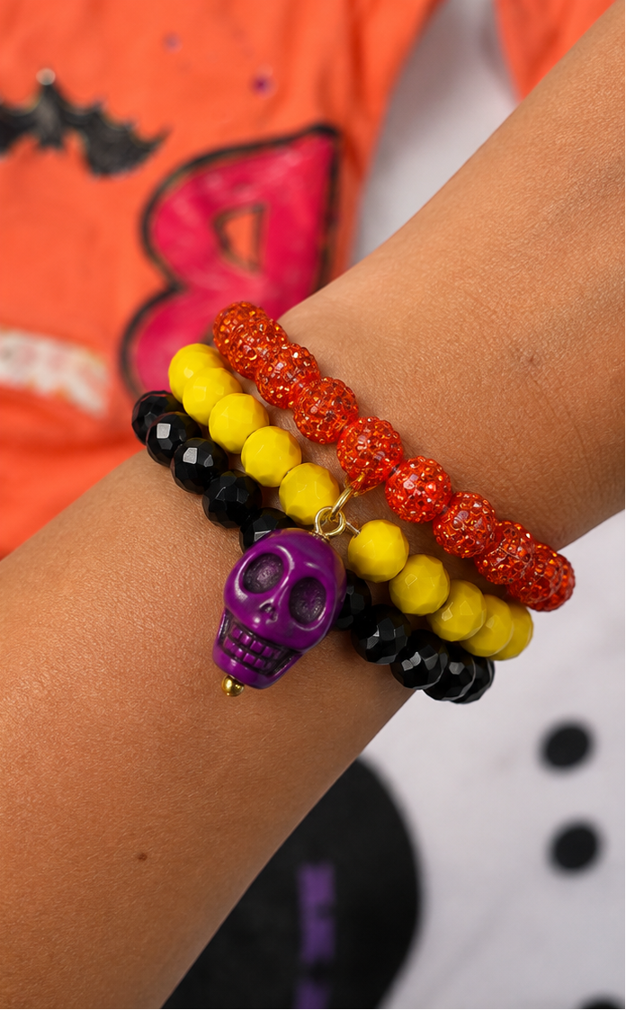 Three beaded stretch bracelets with a purple skull charm, shown in a split view: one laid flat on a white surface and one worn on a person's wrist. The bracelets feature orange glitter beads, yellow beads, and black faceted beads.