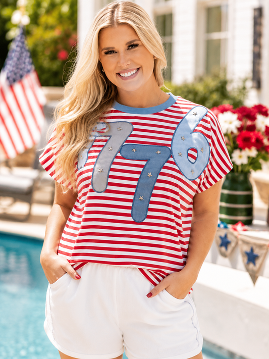 Woman wearing a red and white striped shirt with '1776' and stars, standing by a poolside.