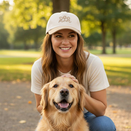 Woman wearing a cap with 'Dog Mom' logo, sitting with a golden retriever in a park.