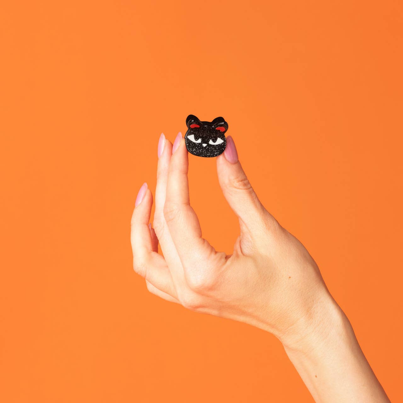 A hand with pink manicured nails holds a small black glittery cat face charm or pin against a solid orange background.