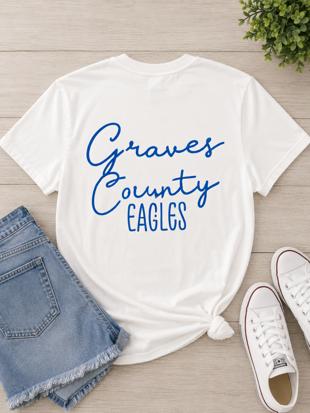 A white short-sleeve T-shirt with 'Graves County EAGLES' printed in blue script across the front, styled in a flat lay with denim shorts, white sneakers, and a small green plant on a light wood background.