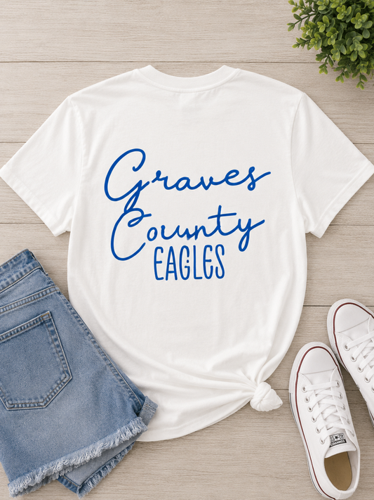 A white short-sleeve T-shirt with 'Graves County EAGLES' printed in blue script across the front, styled in a flat lay with denim shorts, white sneakers, and a small green plant on a light wood background.