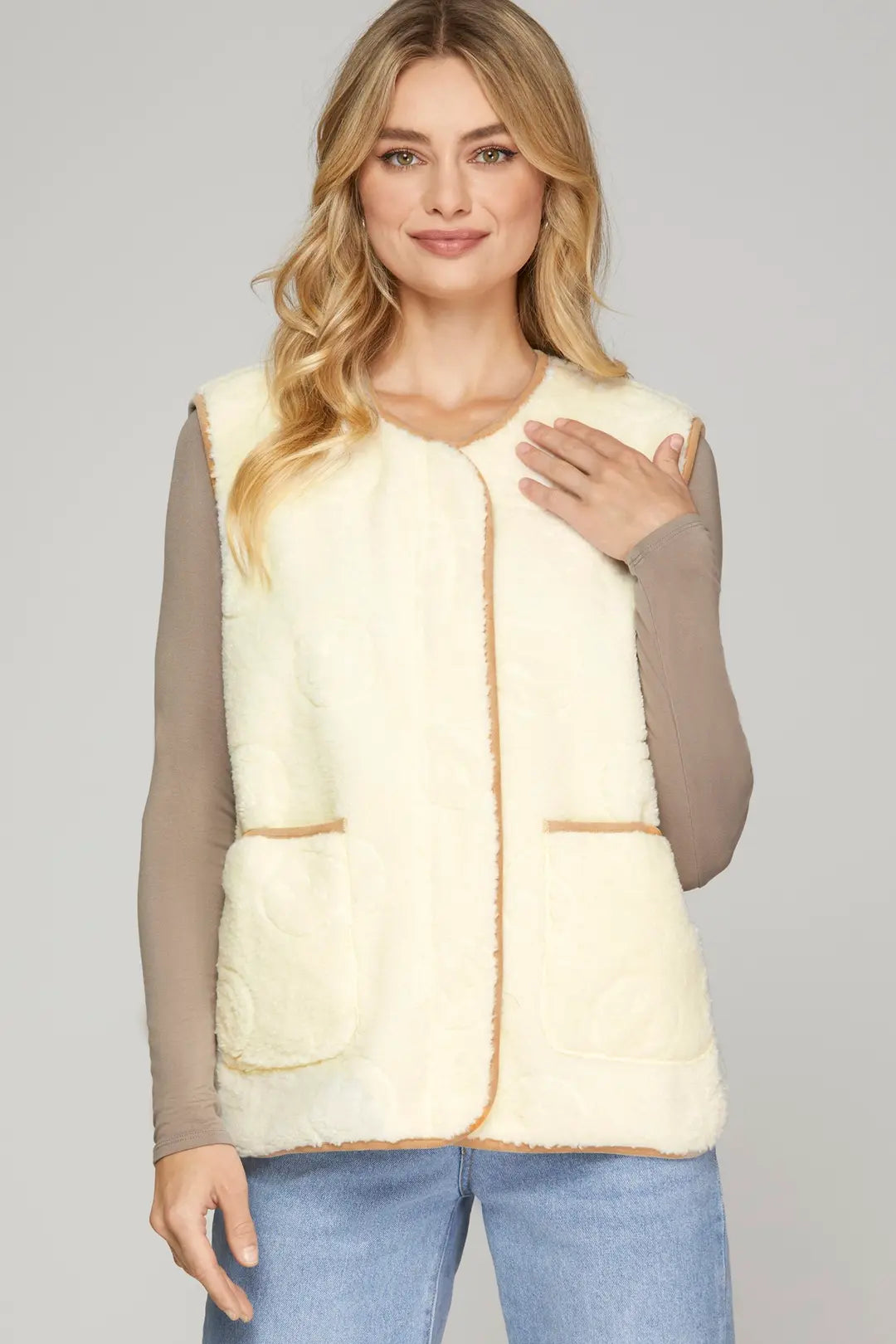 A woman modeling a cream-colored faux fur vest with tan trim over a taupe long-sleeve top and blue jeans, set against a solid light gray background.