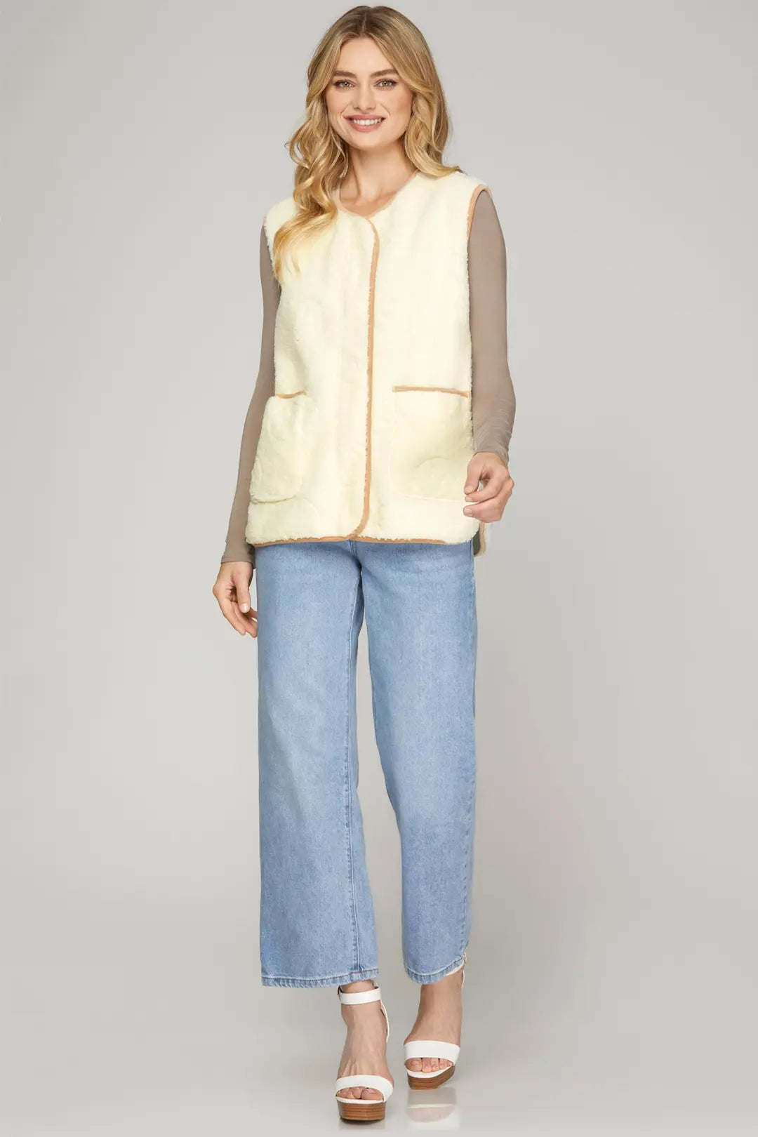A woman modeling a cream-colored faux-shearling vest with tan trim over a gray long-sleeve top and light blue cropped jeans, standing against a light gray studio background.