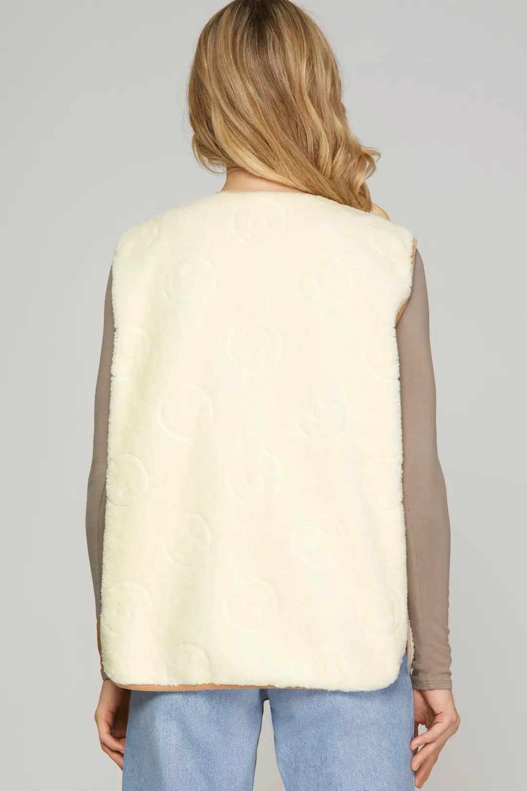 A woman shown from the back wearing a cream-colored faux fur vest with an embossed circular pattern over a taupe long-sleeve top and light blue jeans, against a solid gray studio background.