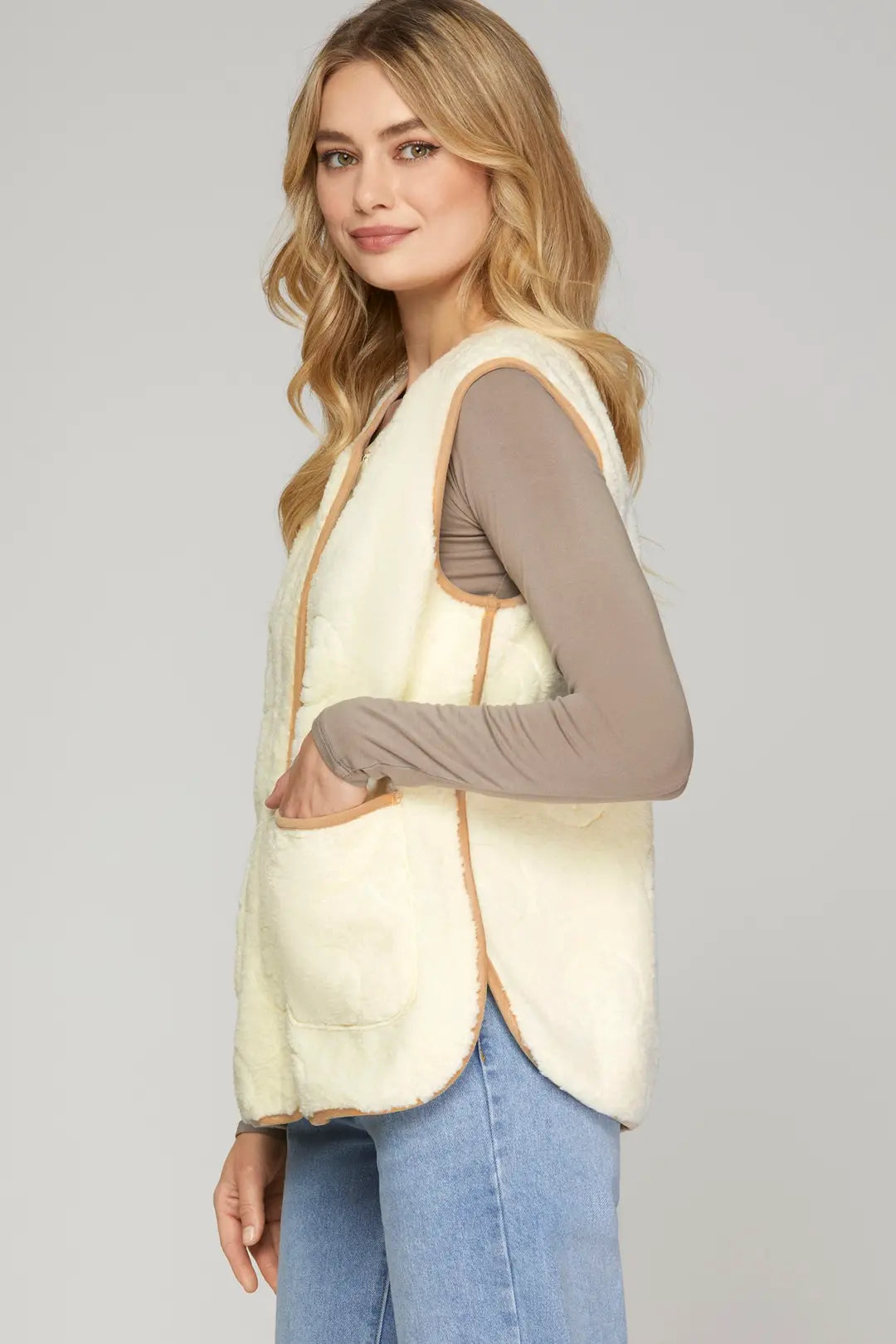 A woman modeling a cream-colored faux-shearling vest with tan trim over a taupe long-sleeve top and blue jeans, photographed against a solid light gray studio background.