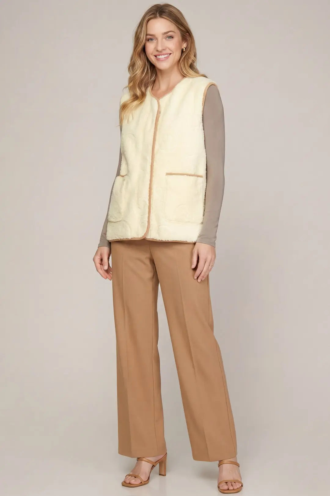 A woman modeling a cream-colored shearling-style sleeveless vest with tan trim over a taupe long-sleeve top and tan wide-leg pants, standing against a neutral light gray studio background.