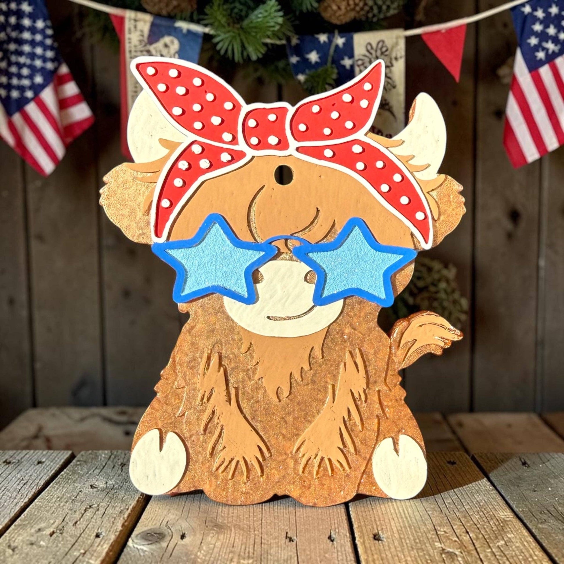 Wooden cow decoration with a red bandana and blue sunglasses on a wooden surface with American flags in the background.
