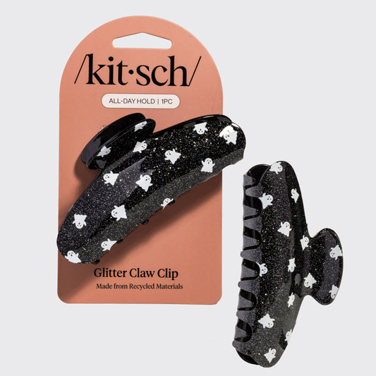 A black glitter claw clip with a white ghost pattern, shown next to its peach-colored retail packaging card by kit sch.