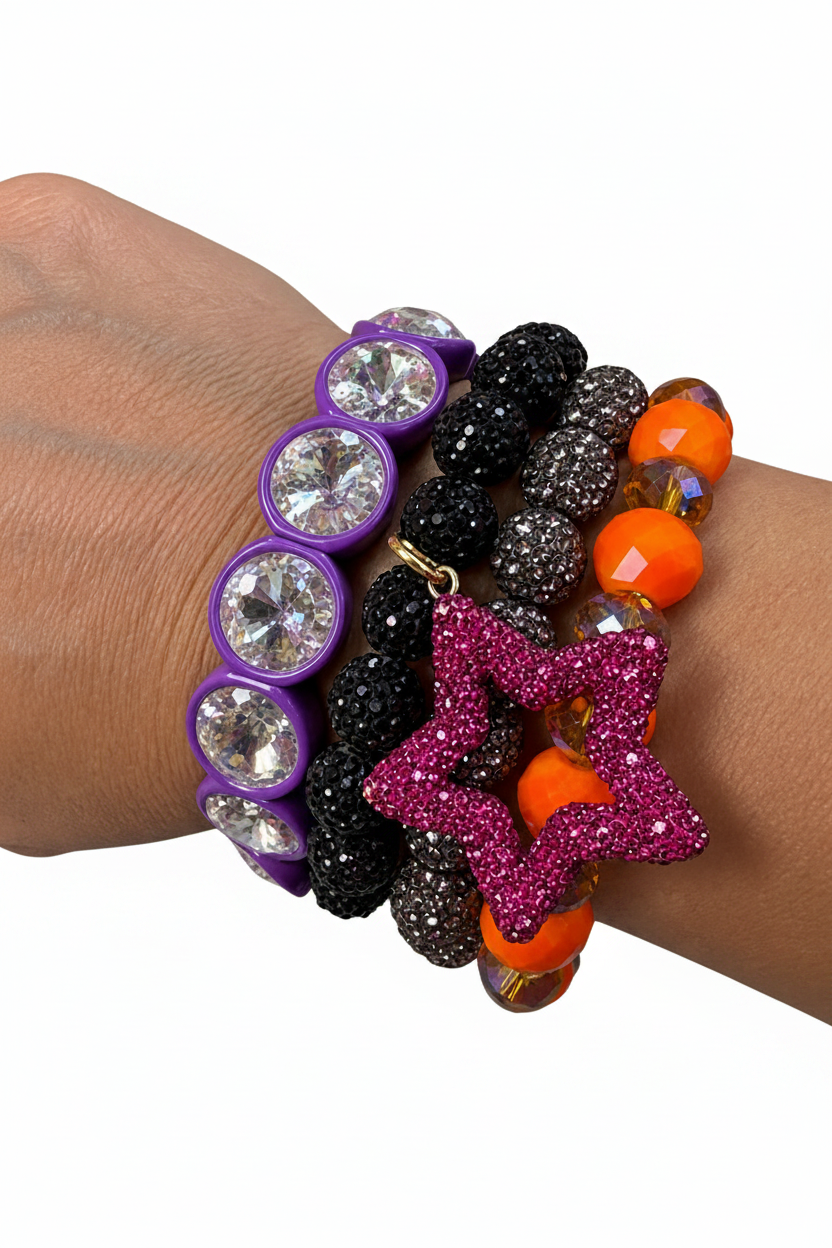 A close-up of a person's wrist wearing four beaded bracelets. The bracelets include purple round bezel-set clear rhinestones, black and smoky gray beaded strands, bright orange faceted beads, and a prominent pink glitter star charm.