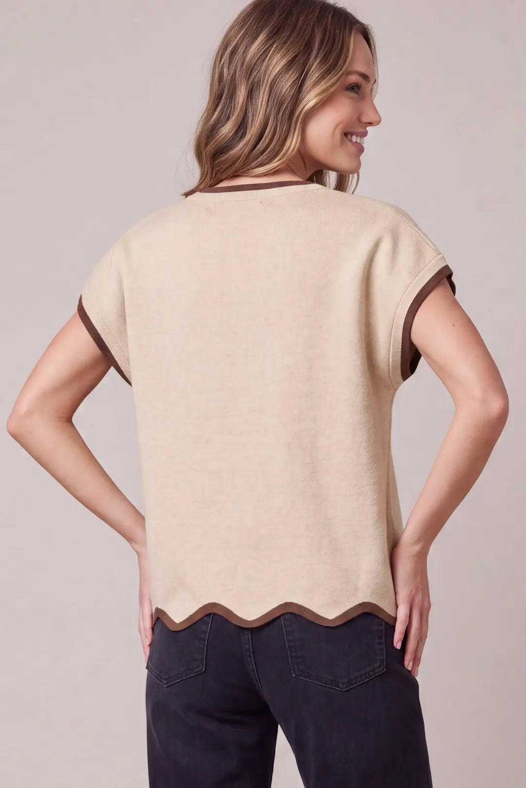A woman shown from the back wearing a light beige short-sleeve knit top with dark brown trim along the neckline, sleeve cuffs, and scalloped hem, paired with dark jeans against a plain light gray studio background.