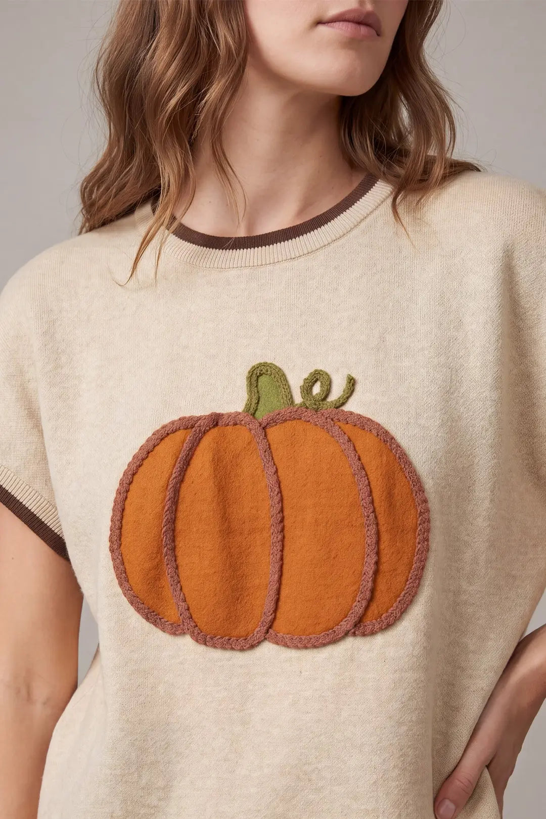 A close-up of a woman wearing a cream-colored knit sweater with a large orange felt pumpkin applique on the front, featuring a green stem and brown ribbed trim.