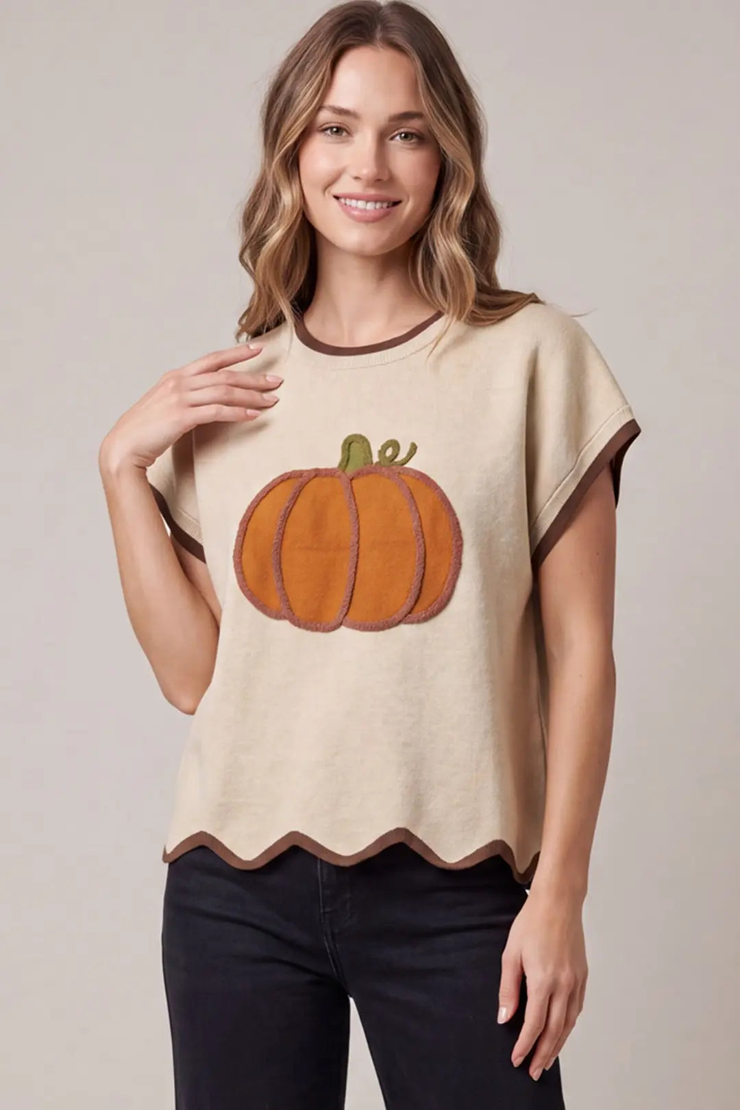 A woman wearing a beige short-sleeve knit top with a large orange pumpkin applique on the front, brown trim, and a scalloped hem, paired with black pants, against a neutral studio background.
