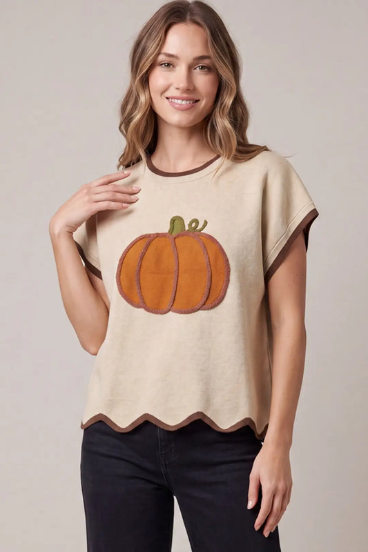 A woman wearing a beige short-sleeve knit top with a large orange pumpkin applique on the front, brown trim, and a scalloped hem, paired with black pants, against a neutral studio background.