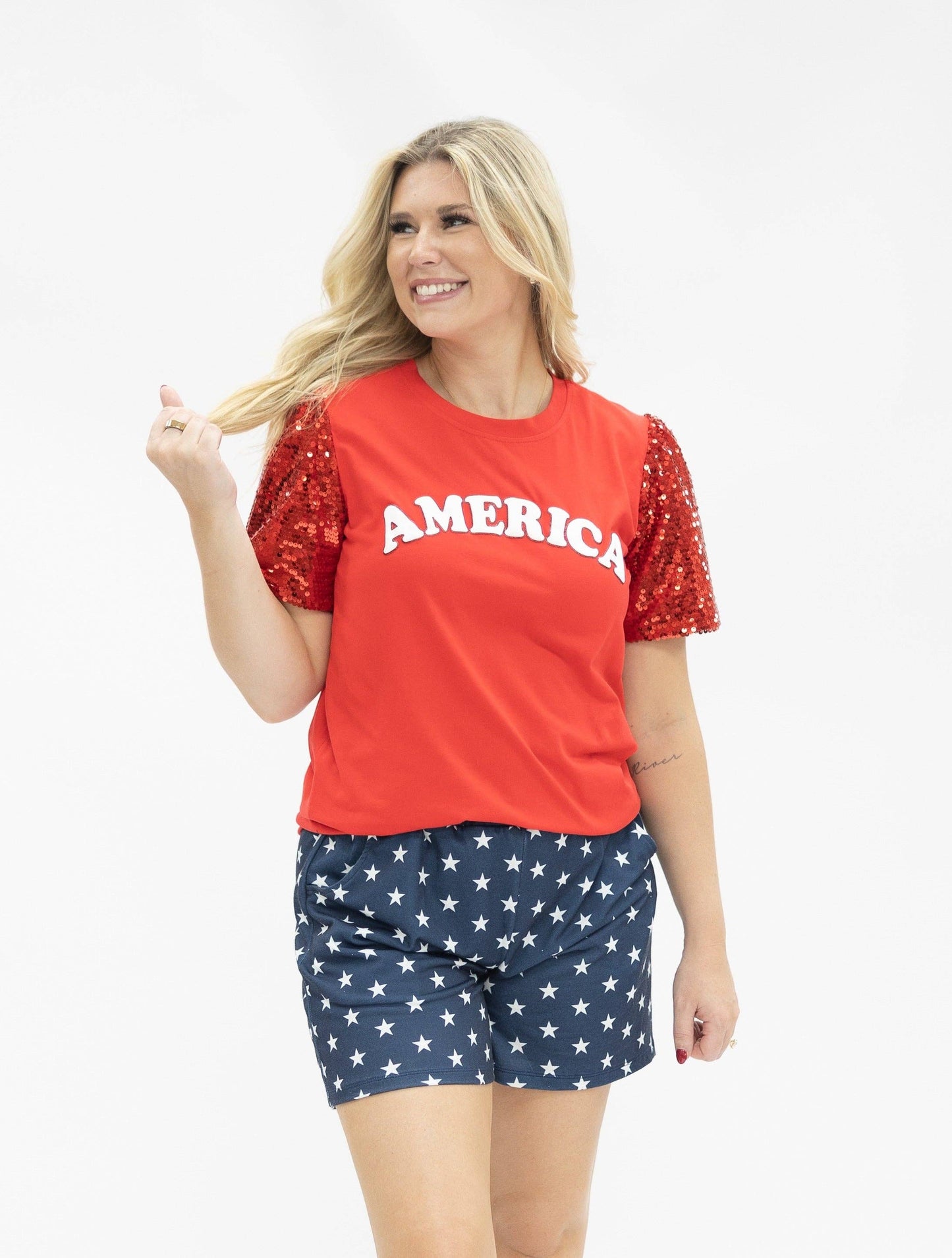Woman wearing a red 'AMERICA' shirt with star sleeves and blue shorts with white stars on a white background