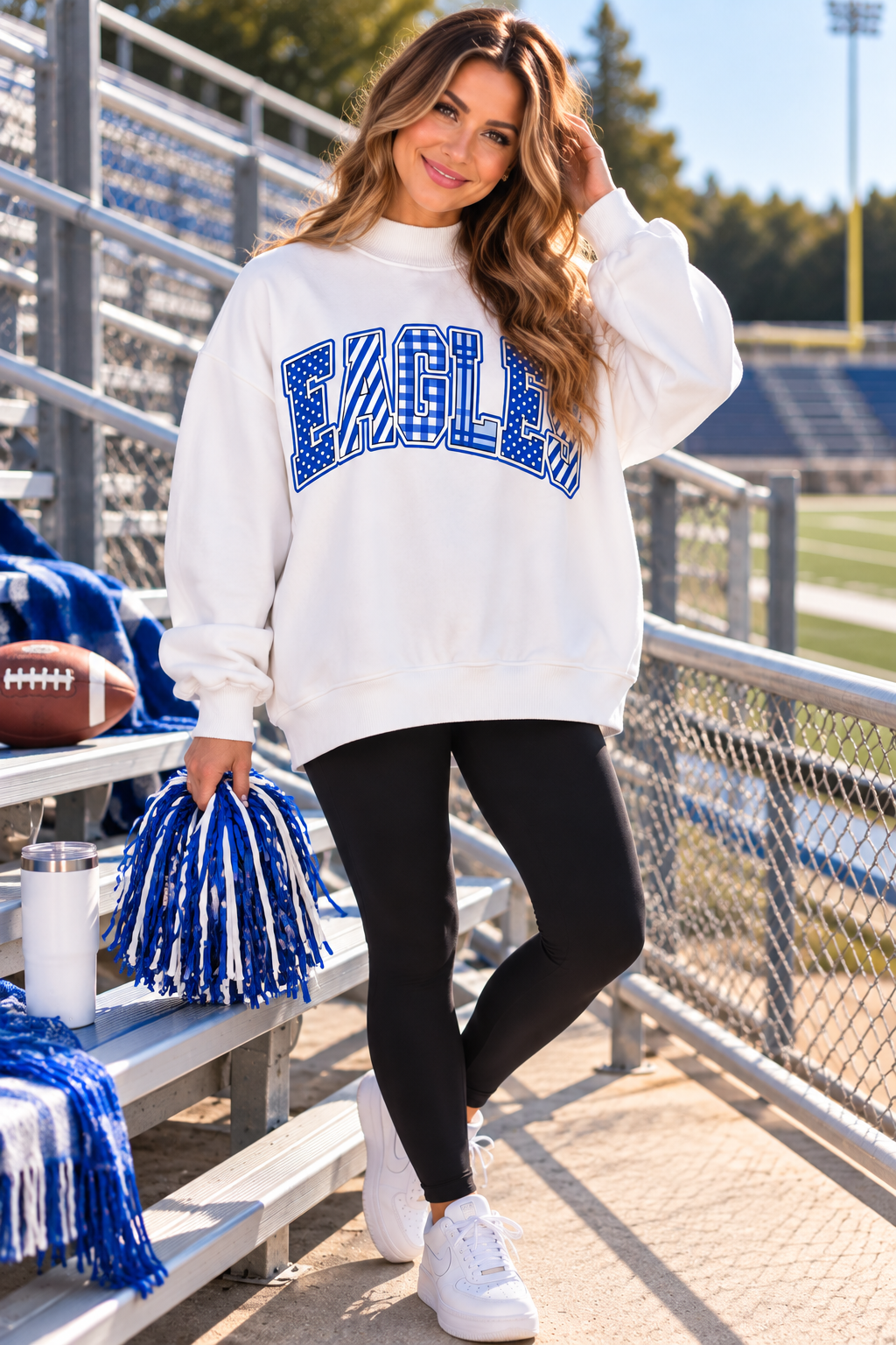 A woman stands on outdoor stadium bleachers wearing a white sweatshirt with a large blue and white 'EAGLES' graphic across the chest, black leggings, and white sneakers. She holds a blue and white pom-pom, with football and bleachers in the background.