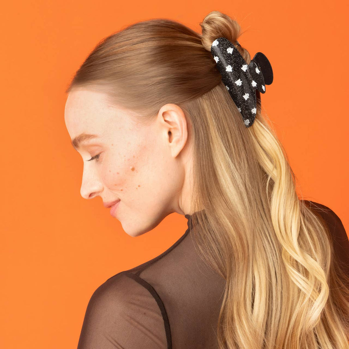 A woman with blonde hair in a half-up bun wearing a black sequined hair claw clip with white star details, set against a solid orange background.