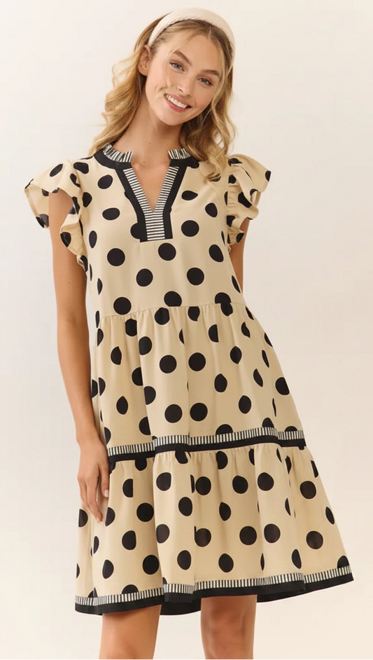 Woman wearing a cream tiered polka dot dress with black dots, flutter sleeves, and contrast striped trim detailing.