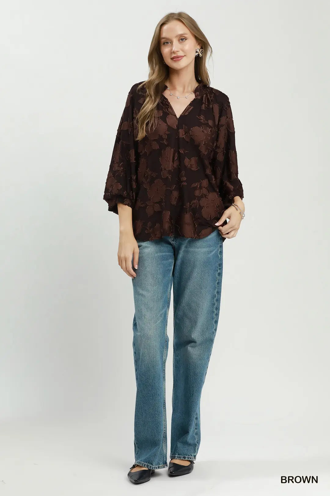A woman modeling a brown floral patterned blouse with three-quarter sleeves and blue jeans, standing against a light gray studio background. The word 'BROWN' appears in the bottom right corner.