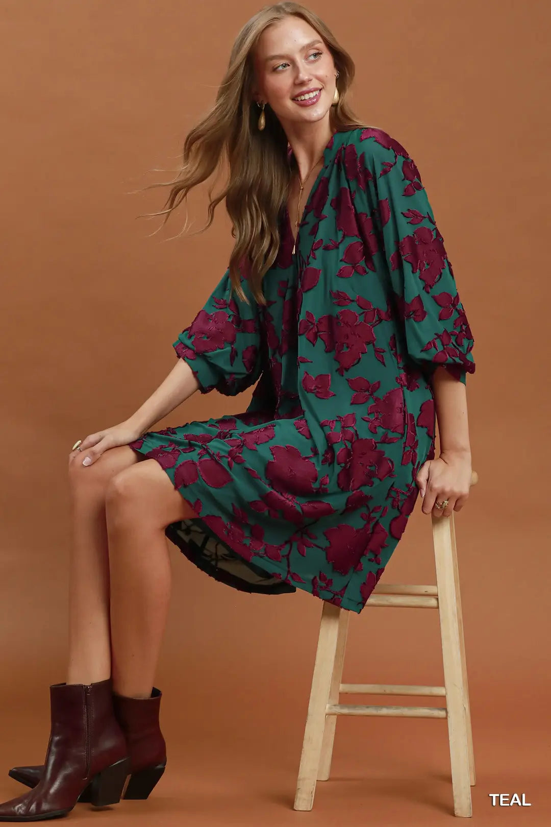 Woman wearing a teal and purple floral dress sitting on a wooden stool against a brown background
